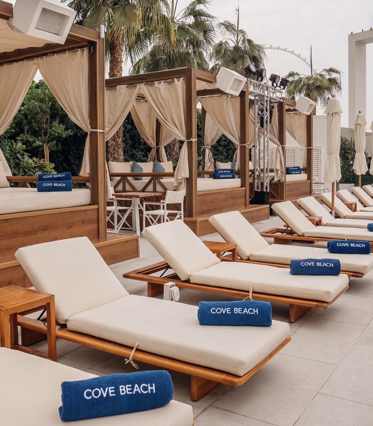 Luxury beach club lounge with wooden cabanas, cream loungers, rolled blue Cove Beach towels, and palm trees in background