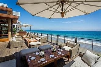 Beachfront restaurant patio with umbrella, lounge seating, dining table, and ocean view under clear blue sky