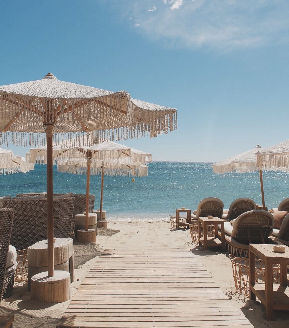 Beach club with beige umbrellas, wooden walkway, lounge chairs, and turquoise sea under blue sky