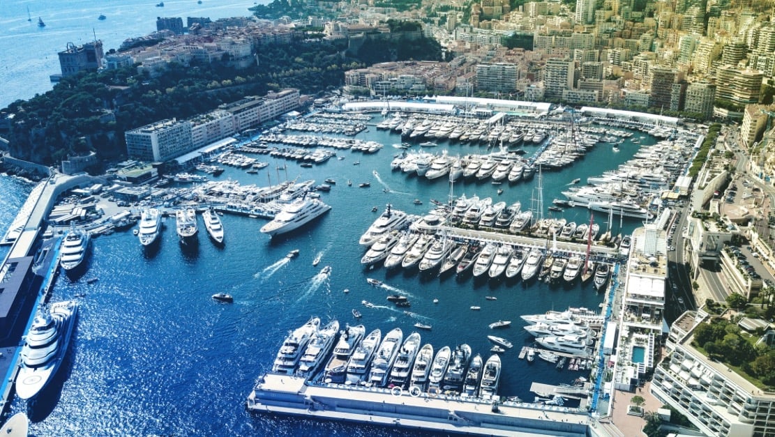 Aerial view of a luxurious marina with numerous white yachts and boats docked in turquoise water, surrounded by coastal buildings and cliffs.