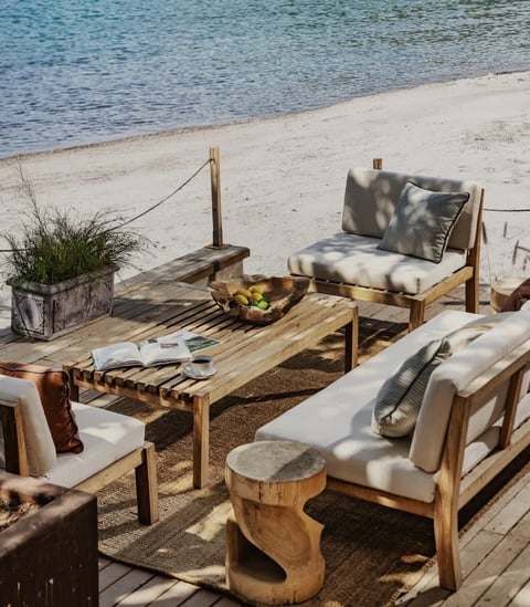 Rustic beach lounge area with wooden furniture, cushioned seating, and potted plants overlooking blue water and sandy shore