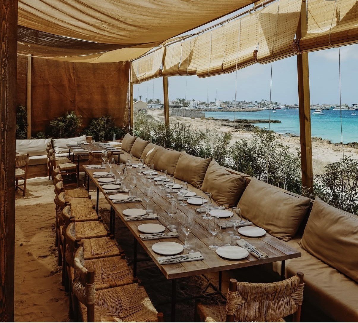 Upscale beachfront dining terrace with long table, tan cushioned seating, woven chairs, and panoramic ocean view through floor-to-ceiling windows