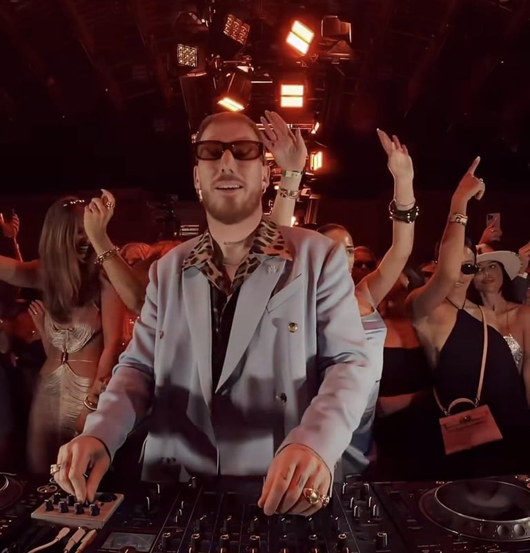 DJ wearing sunglasses and light blue blazer performing at turntables in front of cheering crowd at night event