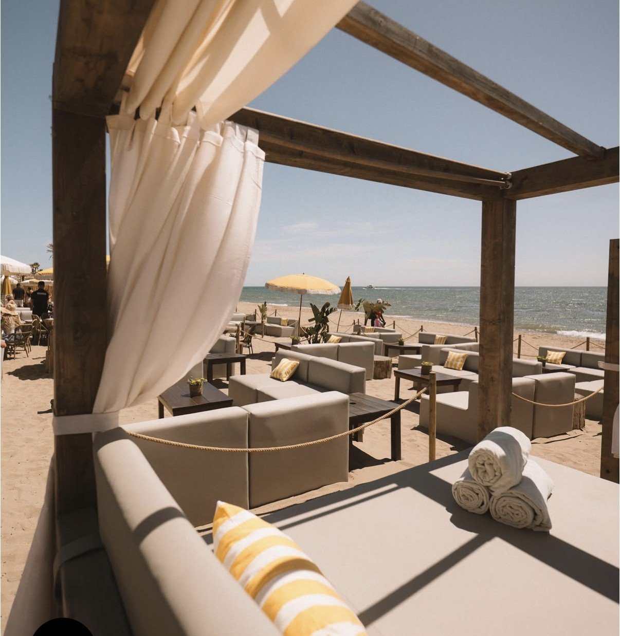 Beach lounge with wooden pergola, white curtains, loungers, and ocean view