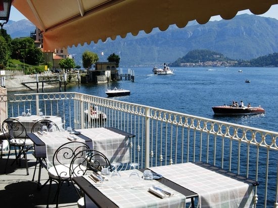 Waterfront dining terrace with white tablecloths, metal railings, and views of a blue lake with mountains and boats