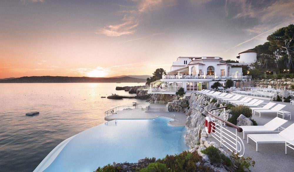 Luxury waterfront resort with infinity pool overlooking the sea at sunset with white buildings and terrace on rocky coastline