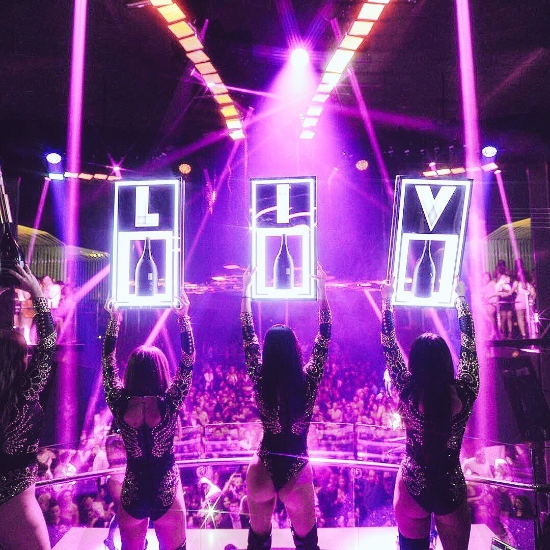 Stage performers on platform surrounded by vibrant purple and pink neon lights with large illuminated letters spelling HEY