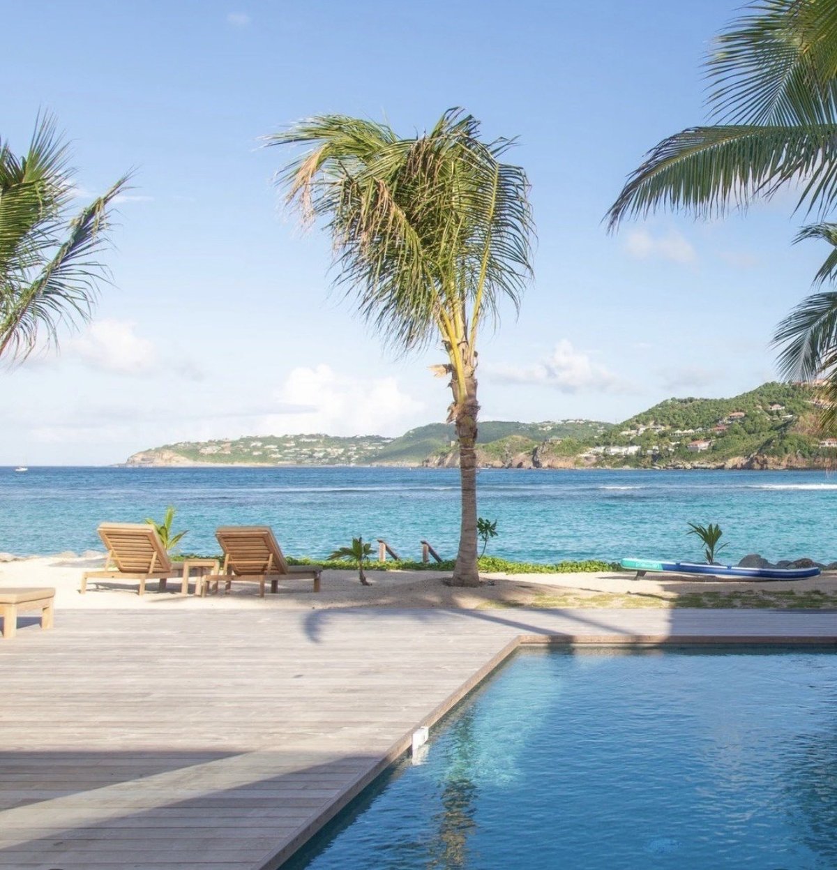 Tropical beachfront resort with wooden deck, lounge chairs, palm tree, infinity pool, and turquoise ocean view with hillside in background