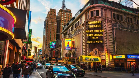 Busy Times Square street with illuminated theater marquees, tall buildings, and pedestrians during daytime