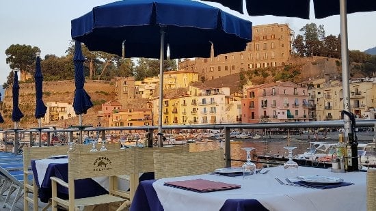 Waterfront restaurant terrace with blue umbrellas overlooking historic coastal town with colorful buildings on hillside