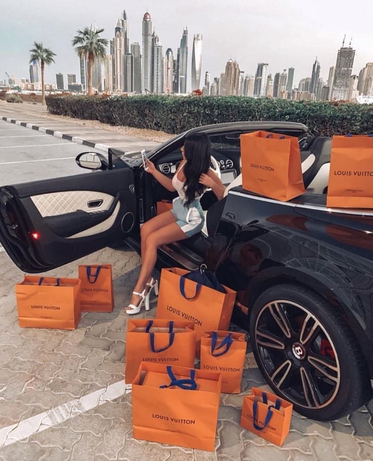 Woman in luxury convertible car surrounded by Louis Vuitton shopping bags with city skyline in background