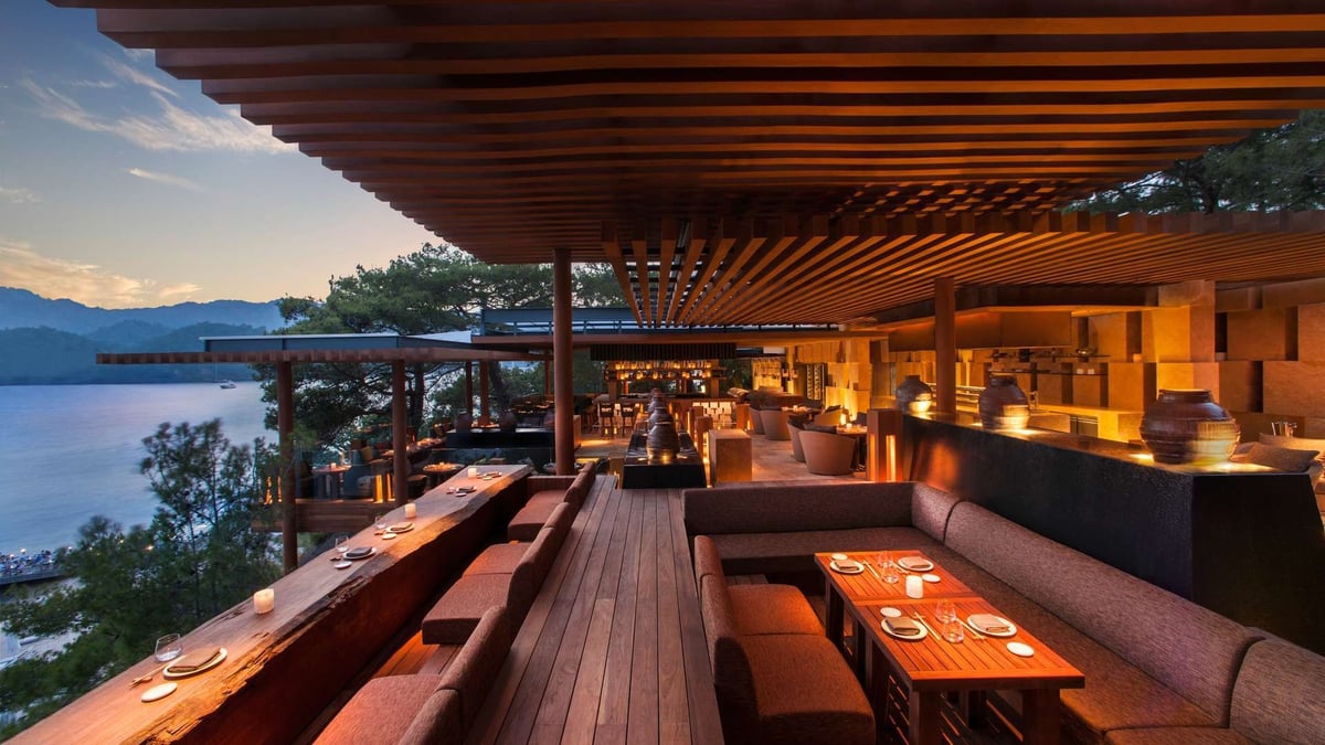 Luxury waterfront restaurant terrace at dusk with wooden deck, lounge seating, and mountain lake views at twilight