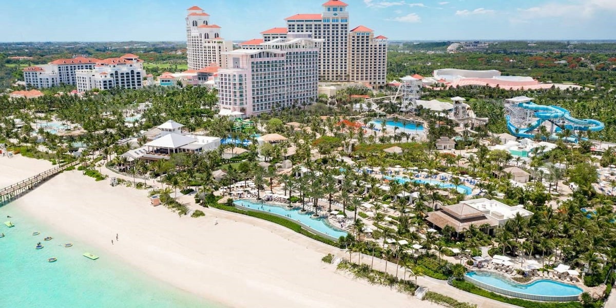 Aerial view of a large beachfront resort with pink hotels, sandy beach, turquoise water, pools, and tropical grounds