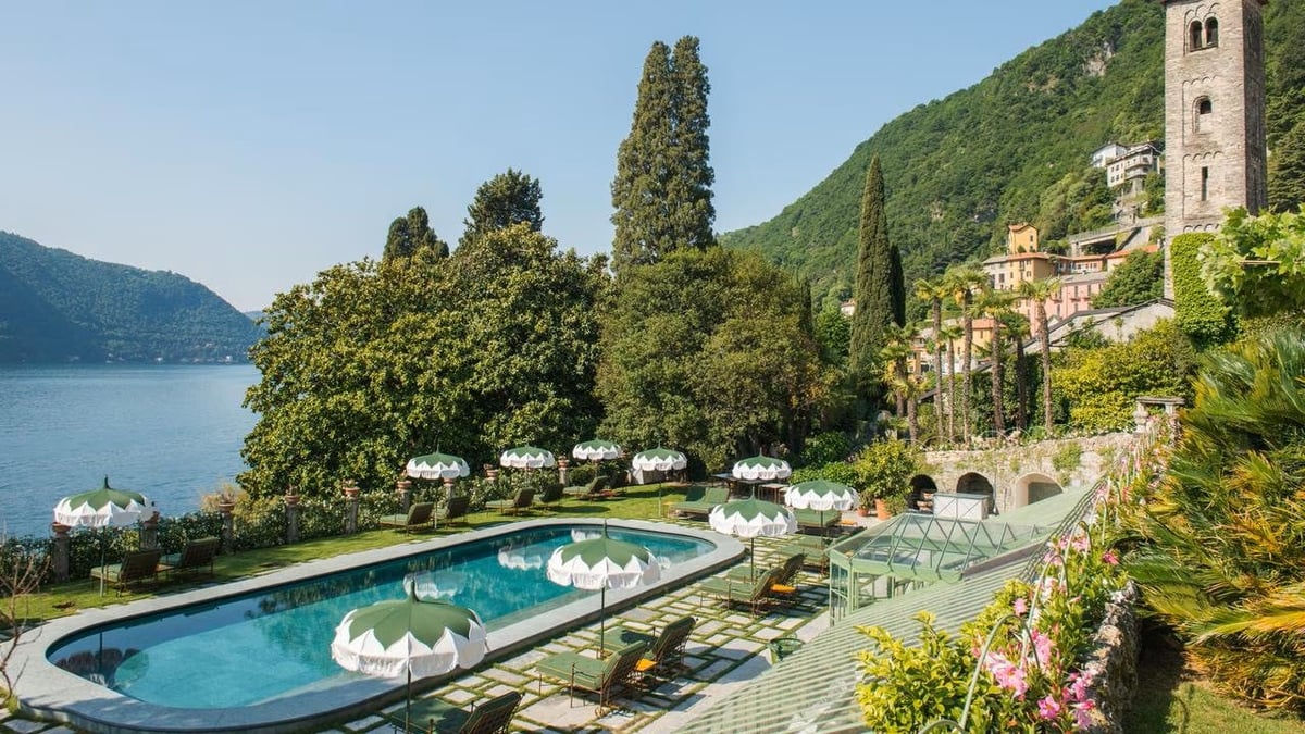 Luxury resort swimming pool overlooking an alpine lake, surrounded by cypress trees and mountains with a historic tower