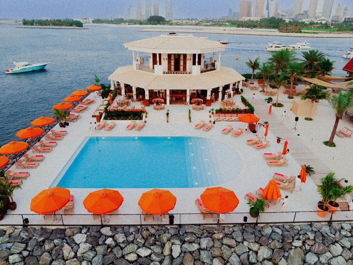 Aerial view of a beachfront resort with rectangular swimming pool, orange umbrellas, palm trees, and octagonal pavilion overlooking blue water and distant city skyline
