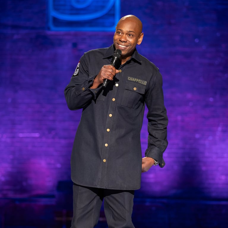 Man performing on stage holding microphone, wearing dark button-up shirt against purple-lit background