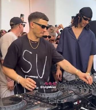Two DJs at turntables wearing sunglasses and dark clothing, performing at an indoor event with spectators in background