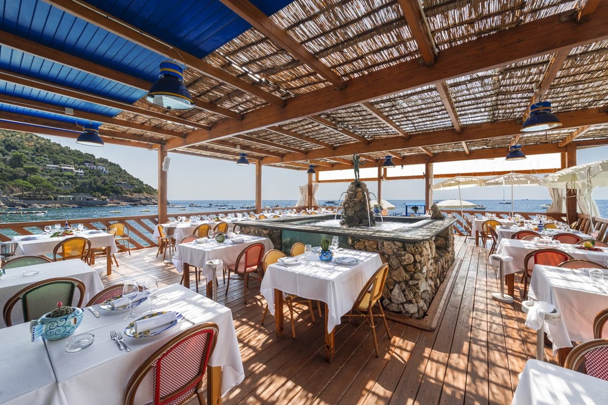 Open-air waterfront restaurant dining area with white tablecloths, wooden beams overhead, and scenic coastal views