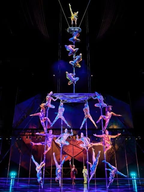 Acrobatic circus performers stacked in pyramid formation under blue lighting with suspended aerial artists above on stage