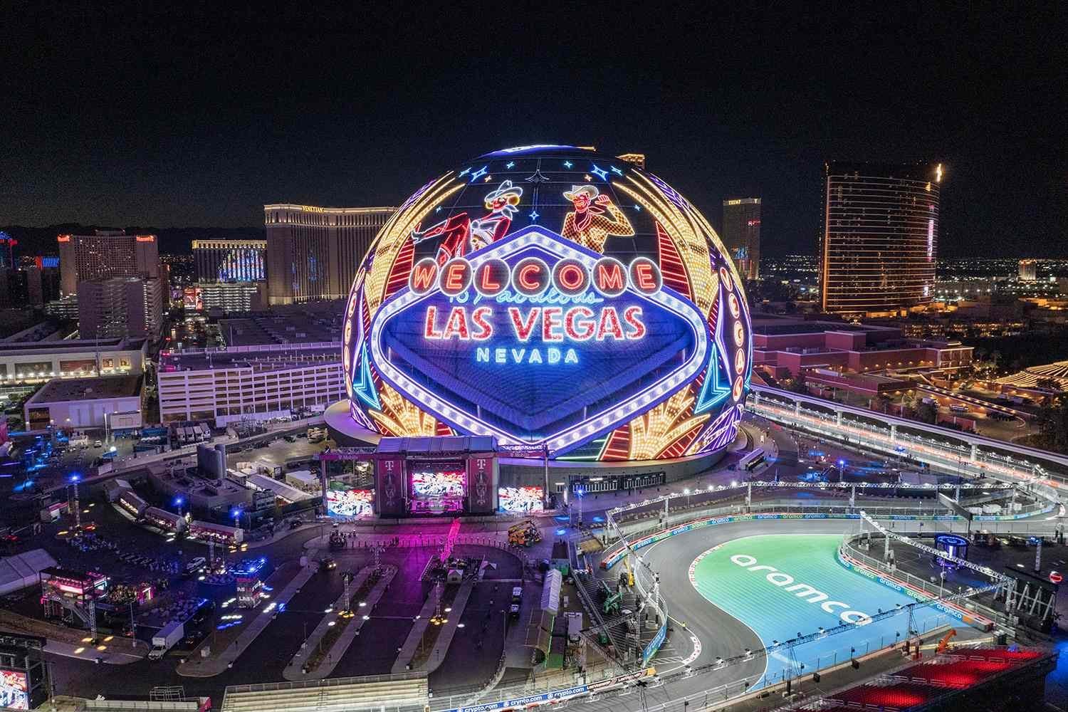 Aerial nighttime view of the Welcome to Las Vegas sign with neon lights above a sports arena, surrounded by city lights and buildings