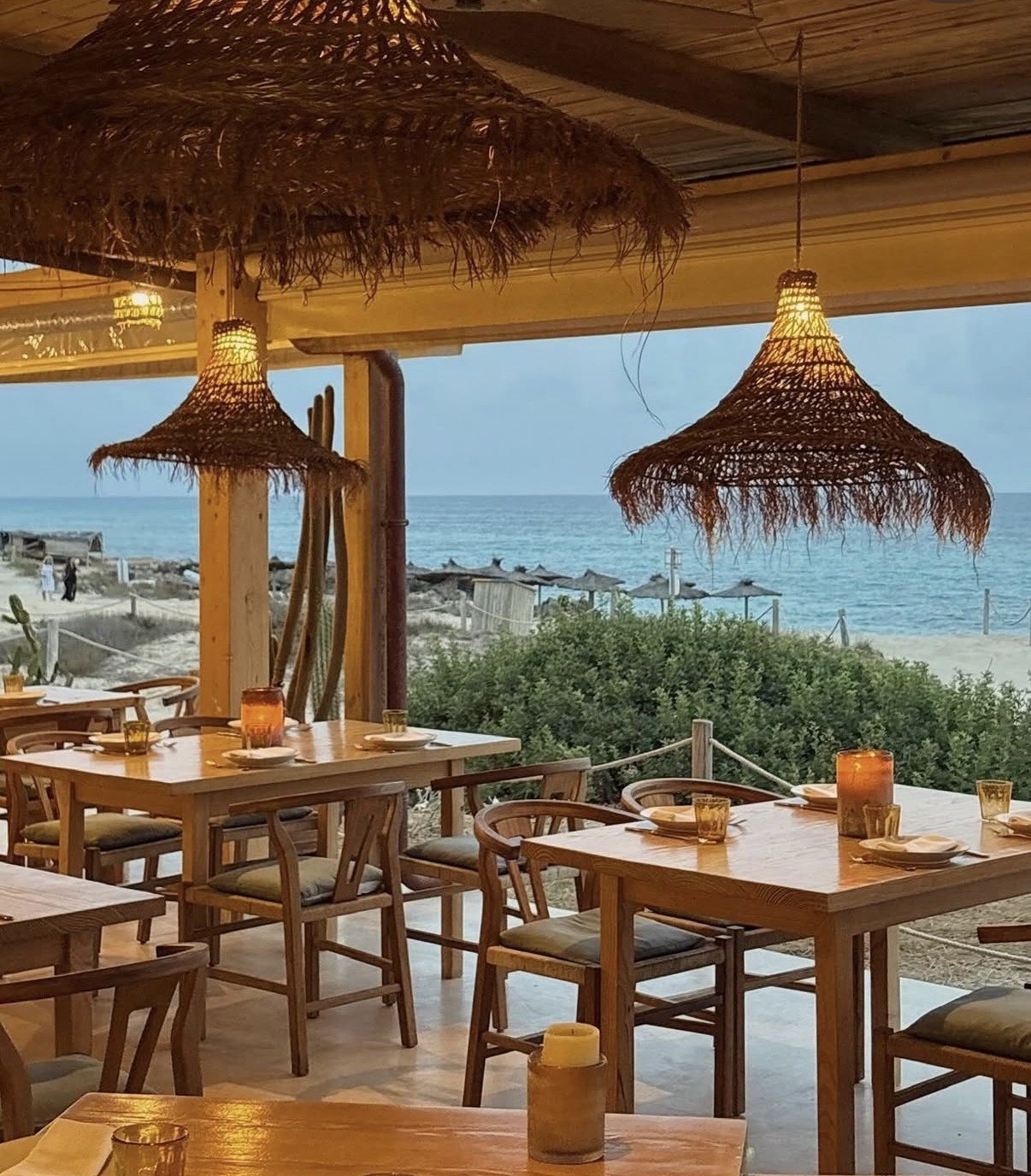 Beachside restaurant terrace with wooden tables, woven pendant lights, and ocean view