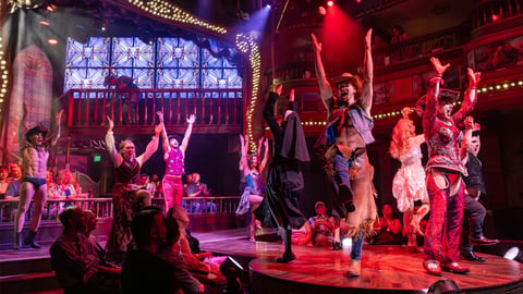 Live stage performance with performers dancing under purple and red lighting, audience watching from foreground in vibrant nightclub setting