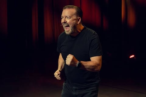 A man in a black t-shirt performs on stage with dramatic red lighting in the background, gesturing expressively while speaking.