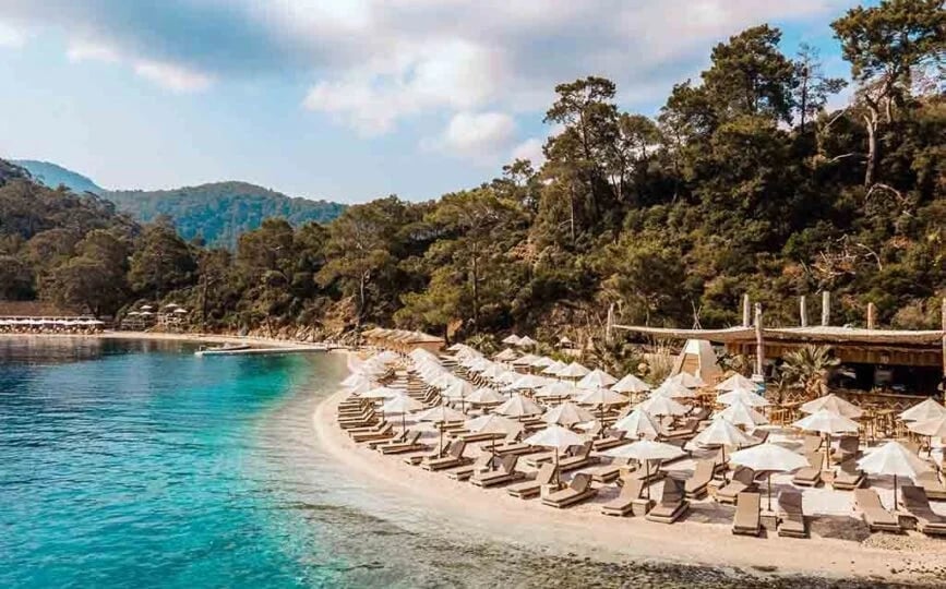 Coastal beach resort with rows of sun loungers and beach chairs on sandy shore beside turquoise water, surrounded by forested mountains