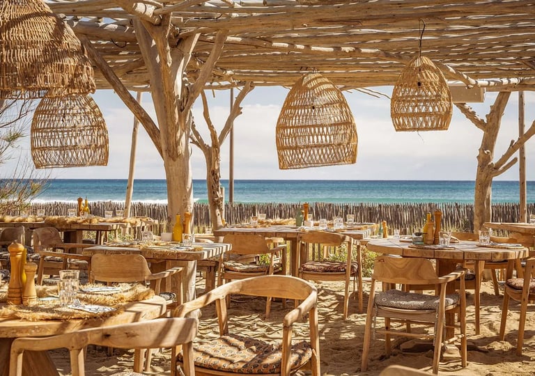 Beachfront restaurant with wooden furniture, woven pendant lamps, and ocean view at sunset.