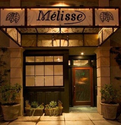 Storefront of Melisse restaurant at night with illuminated signage, black door and windows, flanked by potted plants