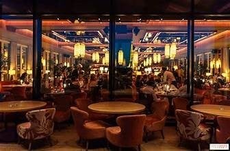 Modern restaurant or lounge interior with round tables, seated guests, warm lighting, and large windows overlooking an illuminated cityscape at night