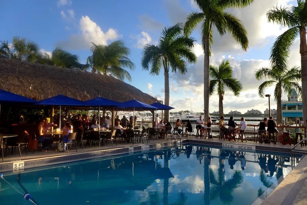 Resort pool area with blue water, palm trees, thatched roof bar, and diners under blue canopy at sunset