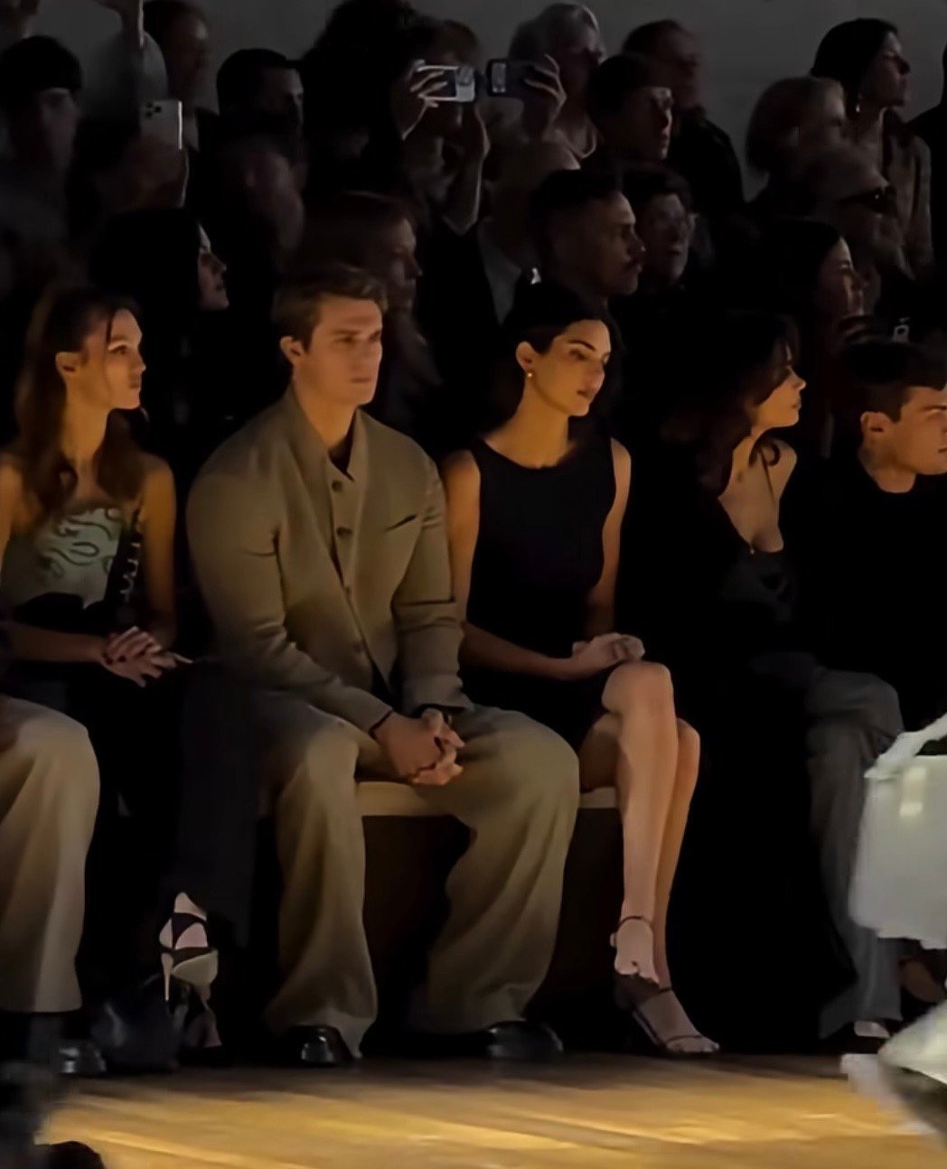 Front row spectators seated at a fashion show, watching models on runway with attentive expressions