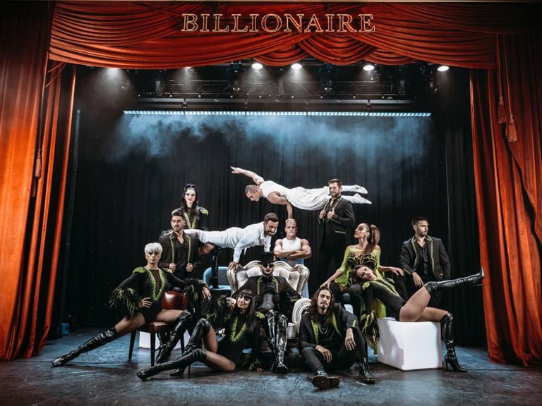 Theatrical cast of BILLIONAIRE posed dramatically on stage with red velvet curtains and dramatic lighting
