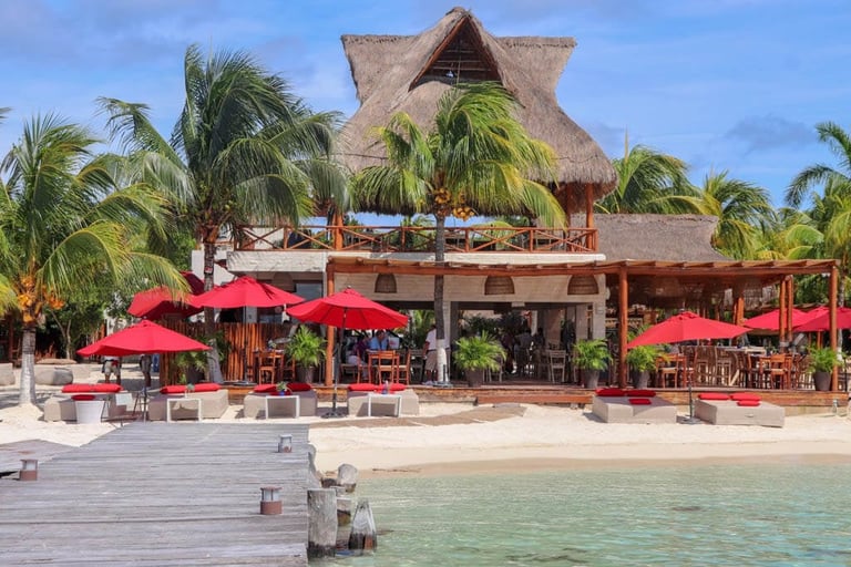 Beachfront resort with thatched-roof buildings, red umbrellas, wooden deck, and palm trees overlooking turquoise water