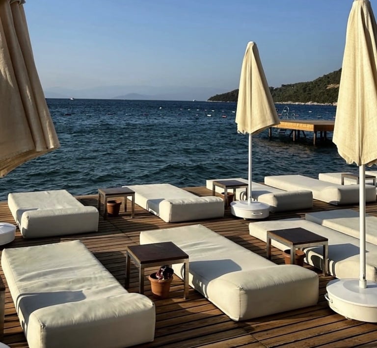 Luxury beachside lounge area with white cushioned sunbeds, cream umbrellas, and wooden deck overlooking turquoise sea and forested coastline