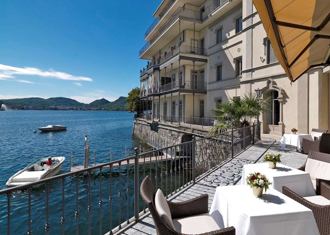 Waterfront terrace with white tables and umbrellas overlooking blue bay with boats and mountains