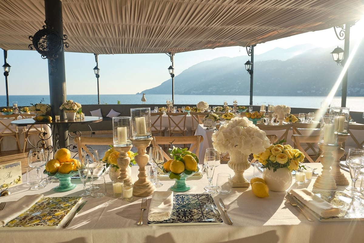 Elegant outdoor dining setup with white tablecloths, yellow flowers, and busts on tables under a striped awning overlooking a coastal lake and mountains