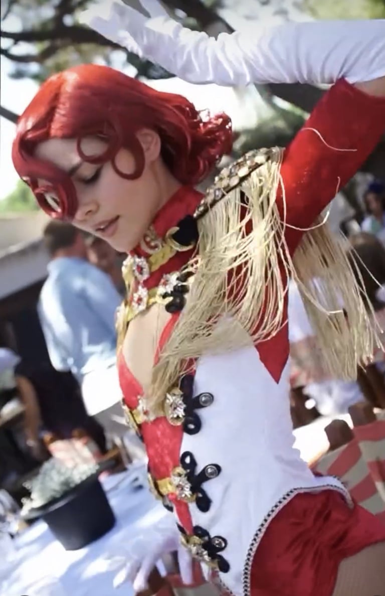 Person in elaborate red and white costume with curled red wig and decorative fringe performing at outdoor event