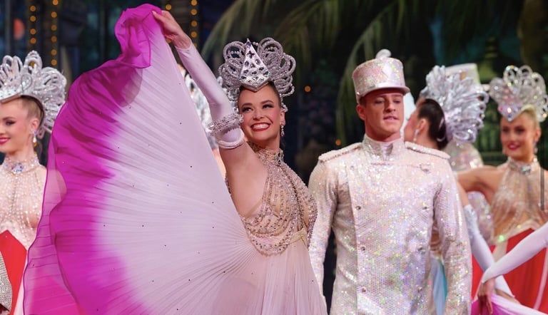 Performers in elaborate costume parade wearing ornate tiaras and shimmering gowns, with vibrant pink fabric flowing in the foreground