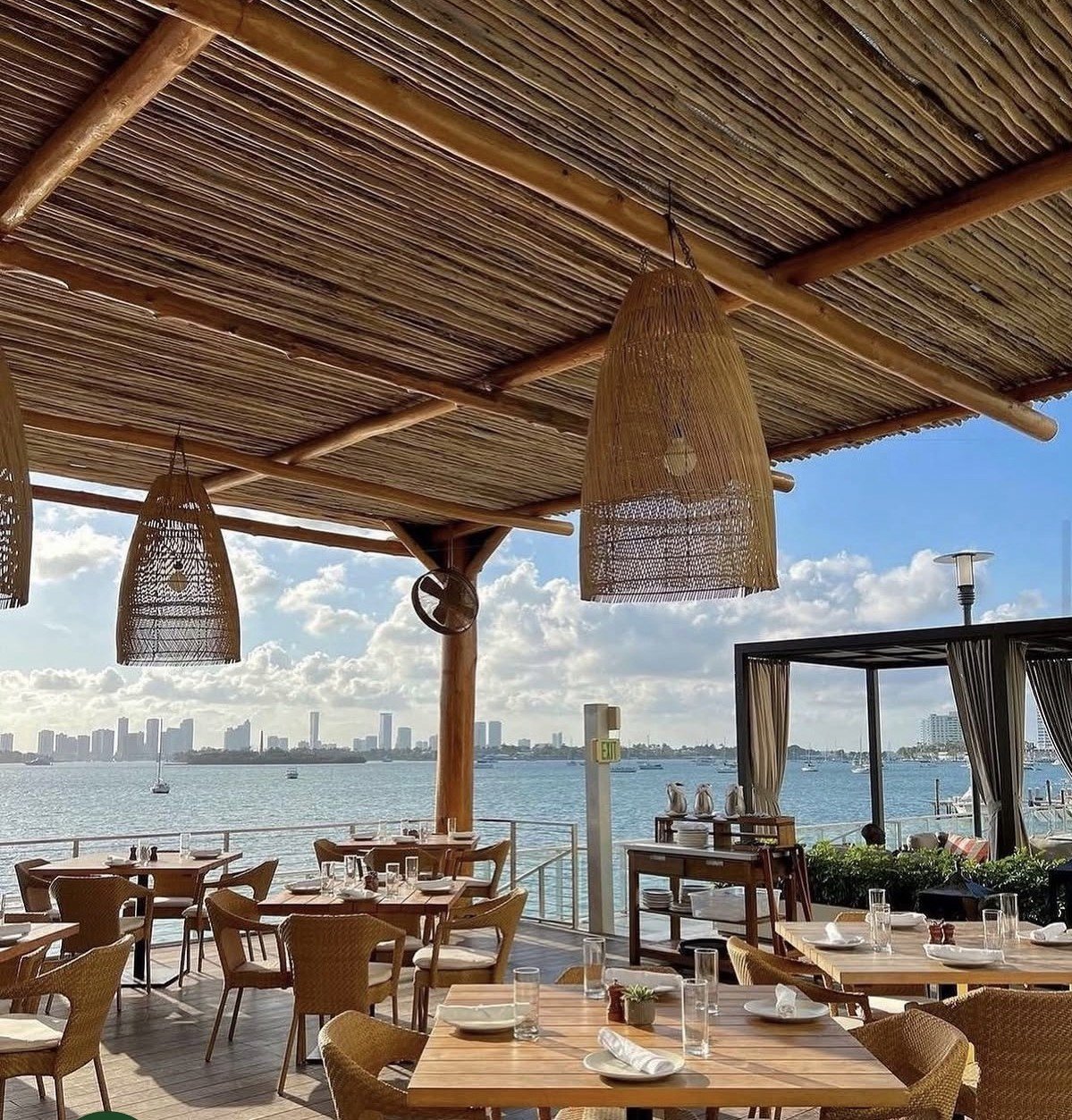 Waterfront restaurant patio with wooden tables, woven pendant lights, and bay views with city skyline in background