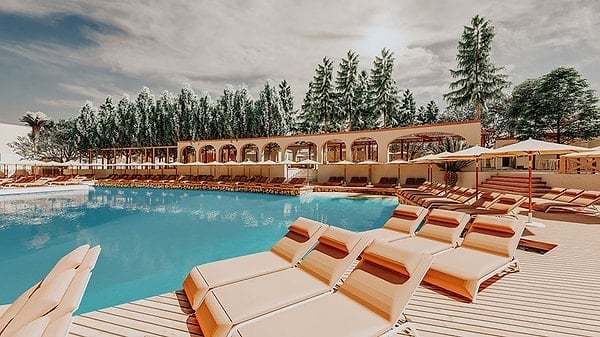 Resort swimming pool with wooden deck chairs, terracotta building, and pine trees under blue sky