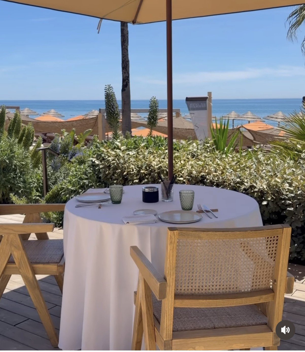 Outdoor dining table with white tablecloth and wooden chairs under a tan umbrella, overlooking a coastal ocean view with Mediterranean landscaping