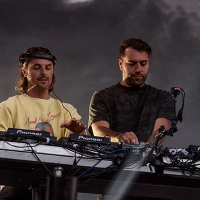 Two DJs performing at turntables in black and white. One adjusts headphones while the other operates the equipment.