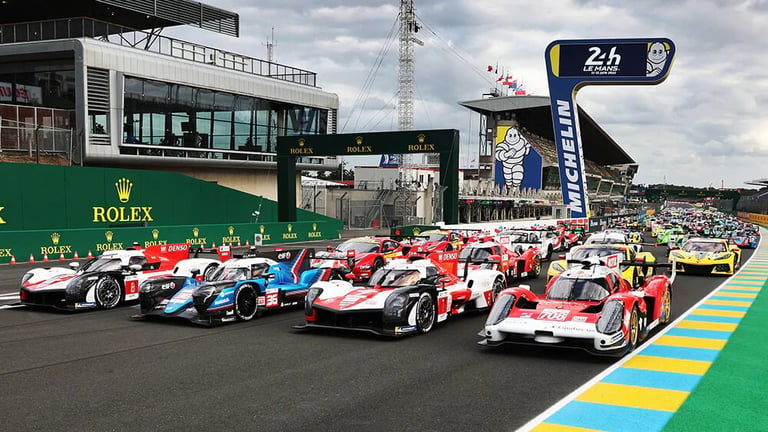 Racing cars lined up on a racetrack at Le Mans, with Rolex signage and pit buildings visible in the background.